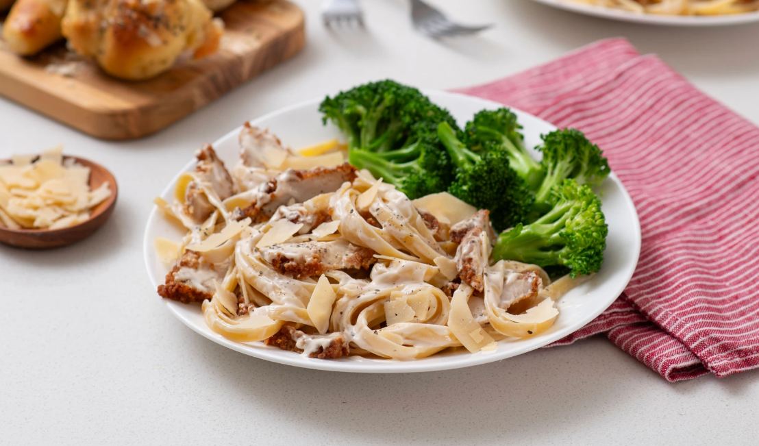 White plate of Chicken Alfredo Pasta with broccoli on top of a red and white striped napkin 
