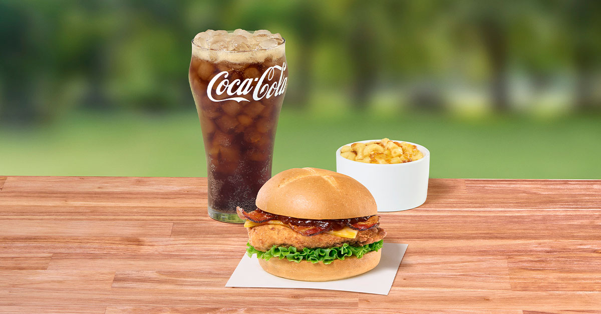 Smokehouse BBQ Bacon Sandwich with Original filet next to a glass of Coca-Cola and a ramekin of Mac & Cheese, sitting on a wooden table outside